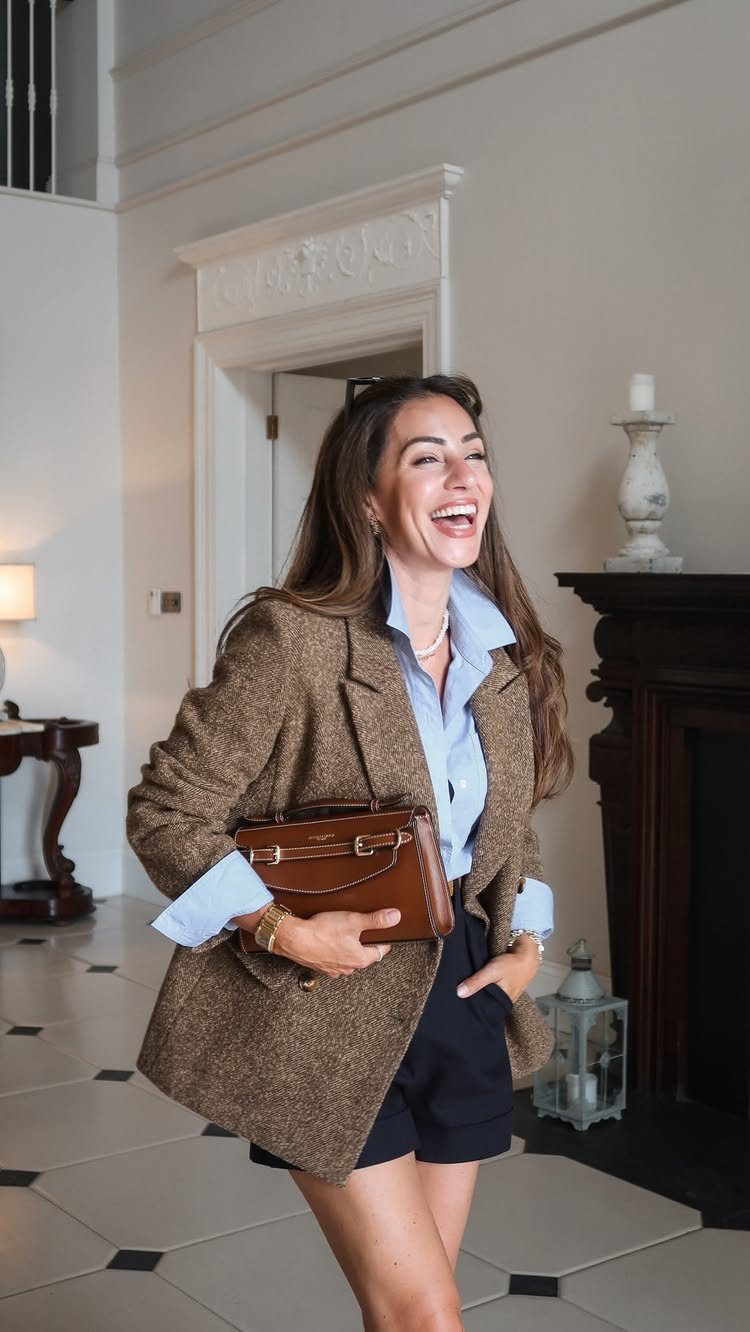 A smiling woman wearing a brown blazer, blue shirt, and dark shorts indoors.