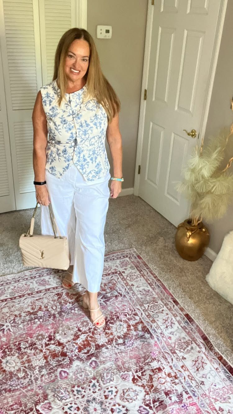 A smiling woman wearing a floral sleeveless top and white trousers standing on a patterned rug.