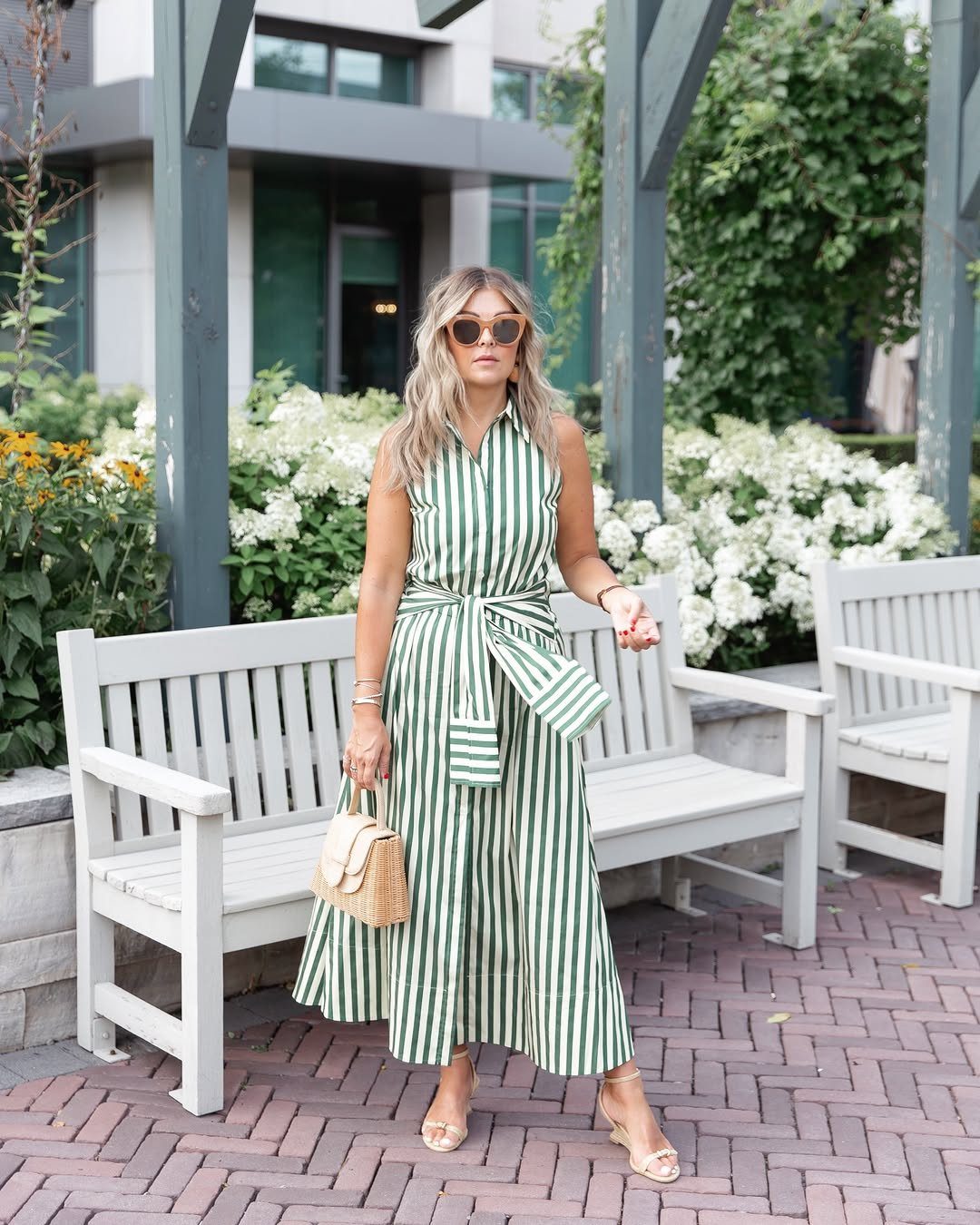 Woman in a green and white striped sleeveless midi dress standing outdoors near white benches.
