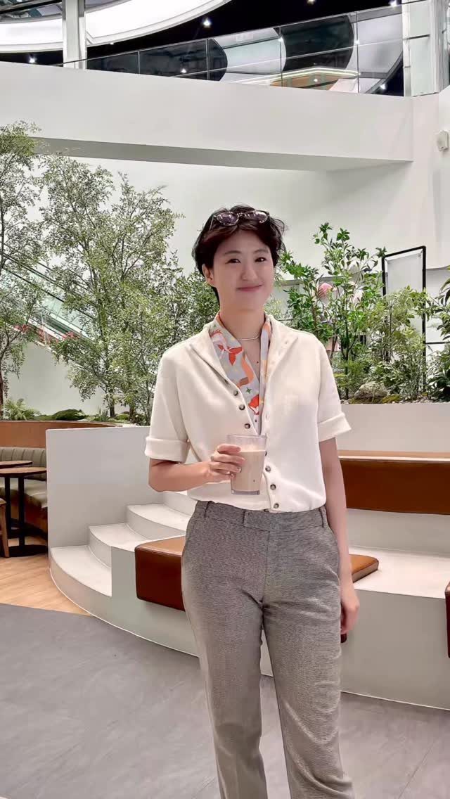 A woman wearing a white cardigan and a patterned scarf while holding a drink indoors.