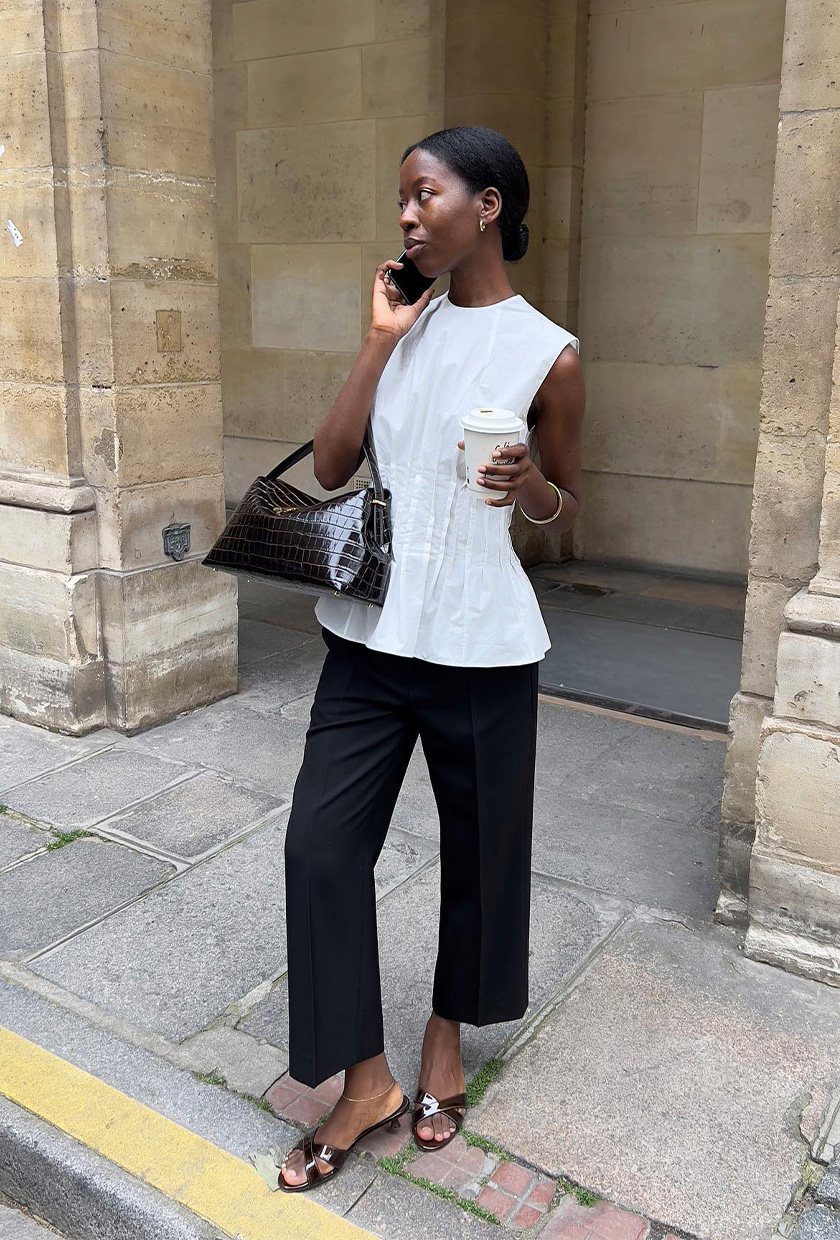 A woman wearing a white sleeveless blouse and black trousers standing by a stone wall.