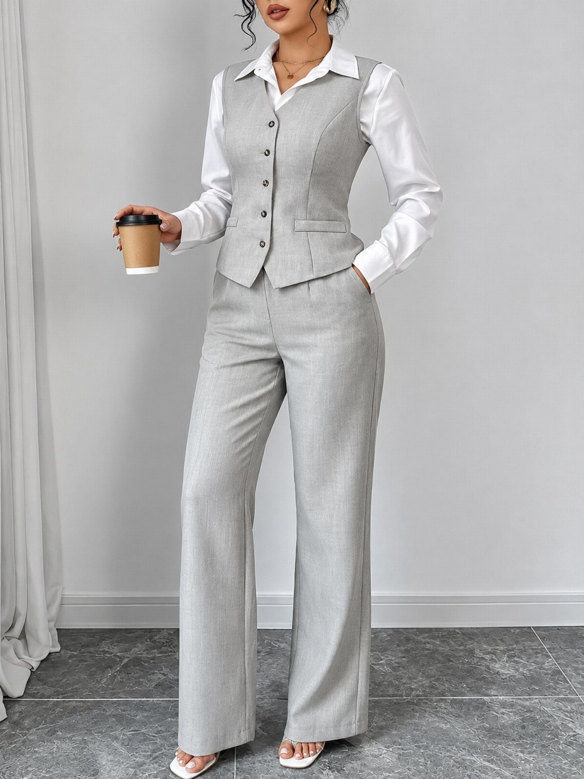 Woman wearing a grey tailored vest, white blouse, and matching wide-leg trousers holding a coffee cup.