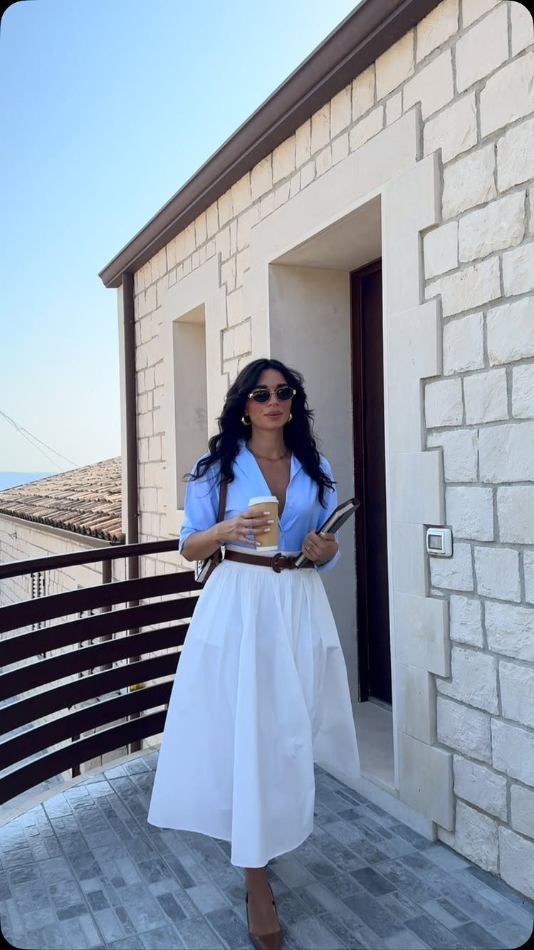 Woman in a blue button-up shirt and white midi skirt standing on a stone balcony.