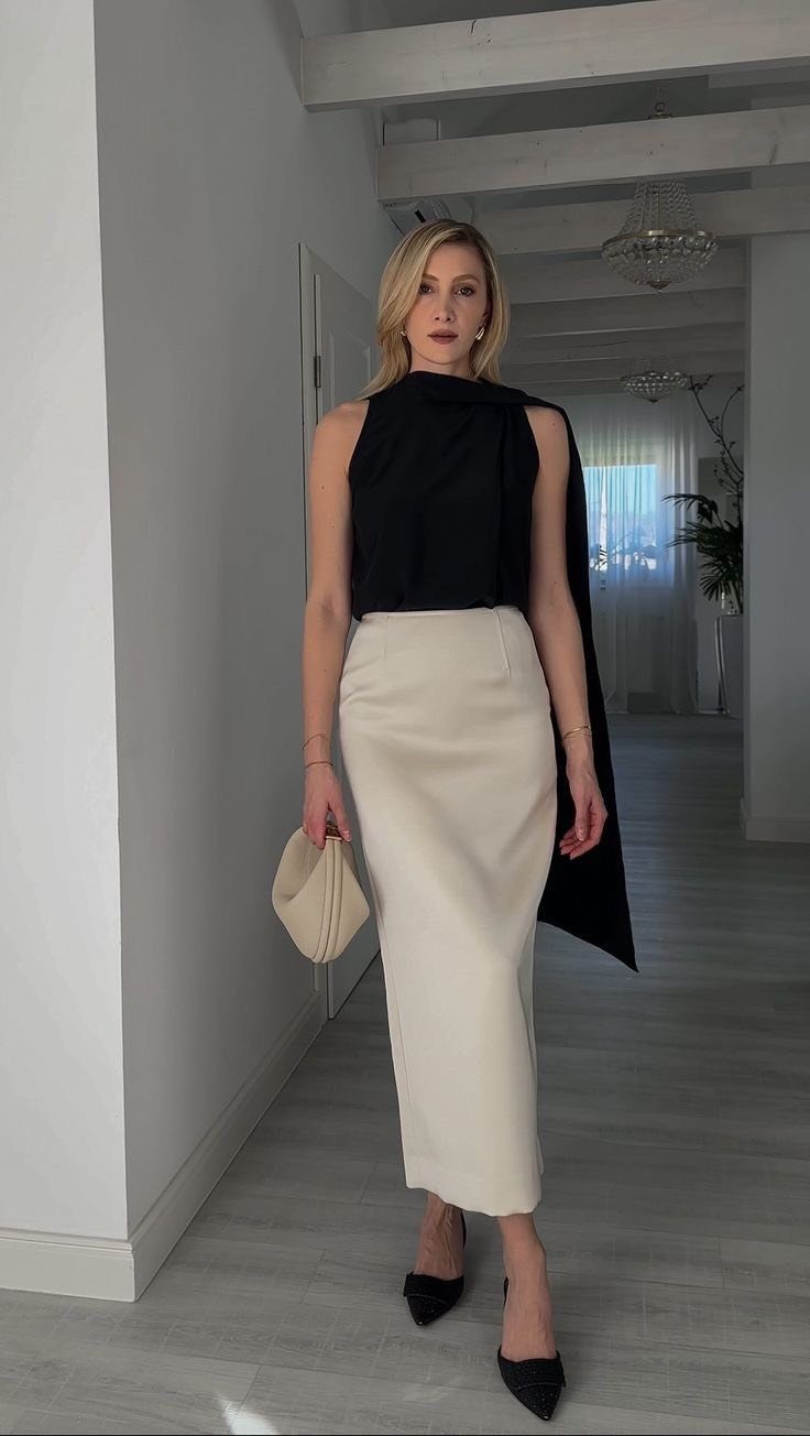Blonde woman wearing a black sleeveless top and cream pencil skirt standing in a hallway.