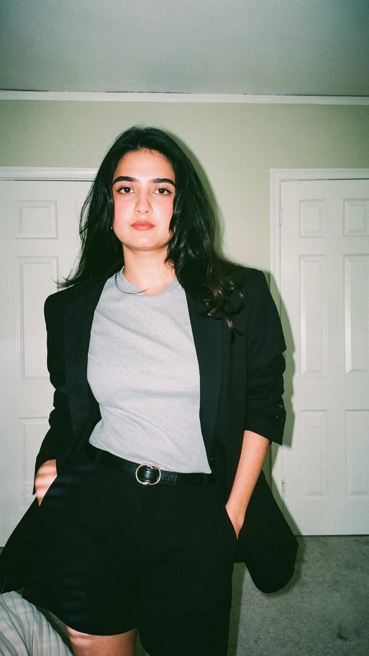 A woman wearing a black blazer, grey t-shirt, and black tailored shorts posing indoors.