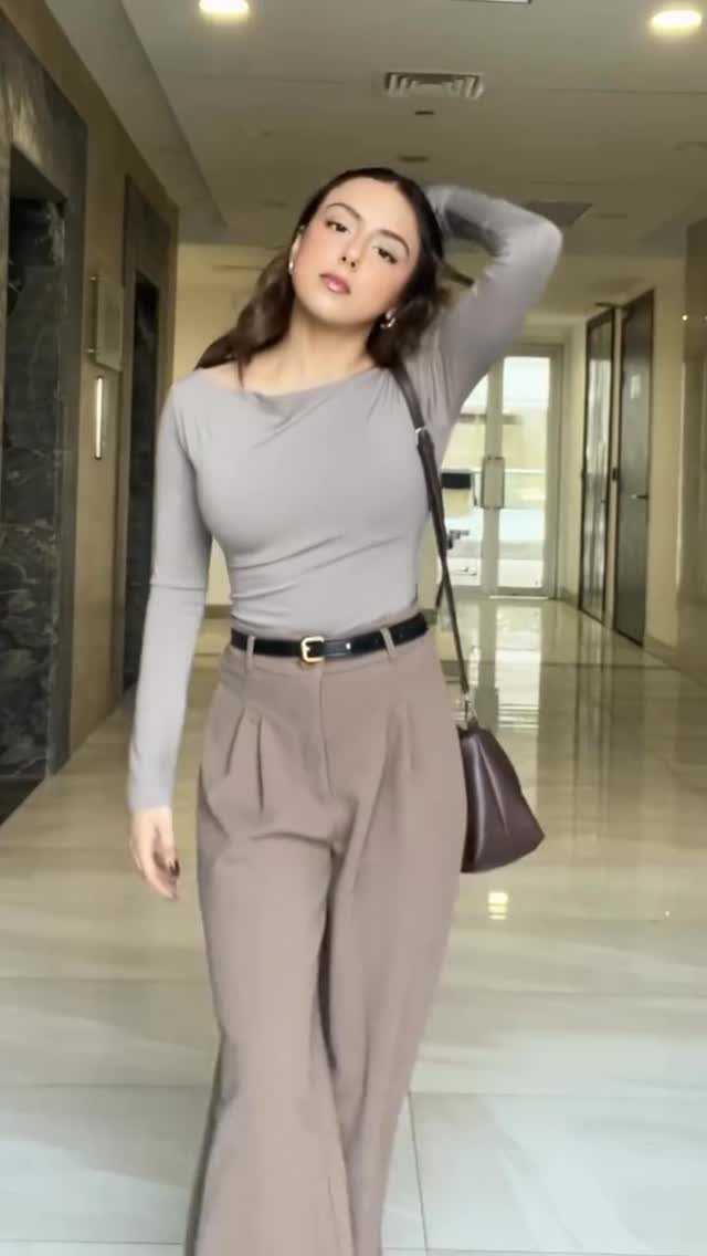 Woman wearing a grey fitted top and tan wide-leg trousers standing in a hallway.