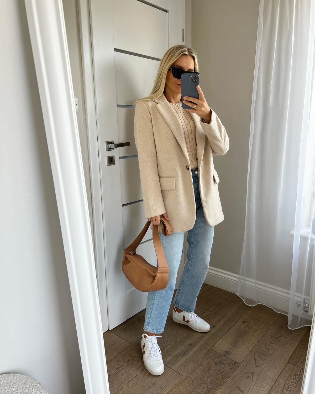 Woman in a beige blazer, cream sweater, and light blue jeans posing for a selfie.