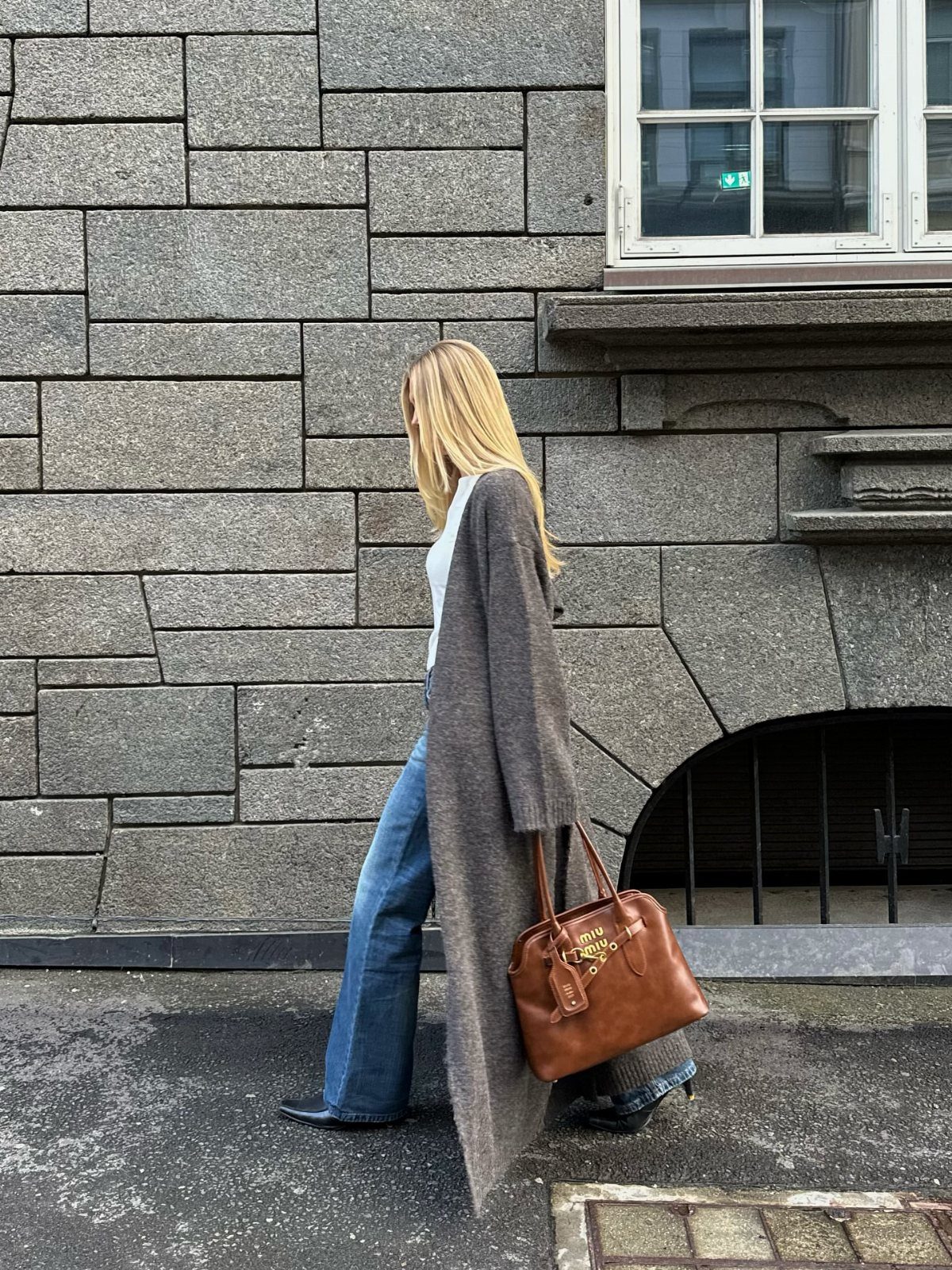 Blonde woman walking in blue jeans, a white top, and a long grey cardigan while carrying a brown handbag.