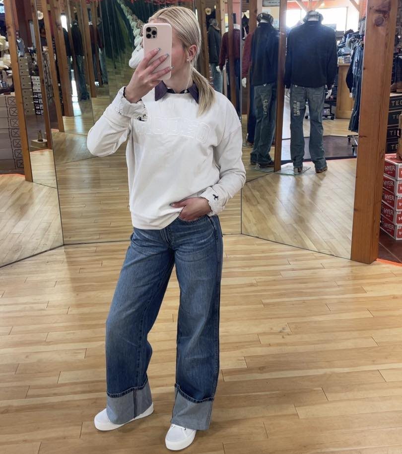 Woman in a white sweatshirt and wide-leg blue jeans posing for a mirror selfie.