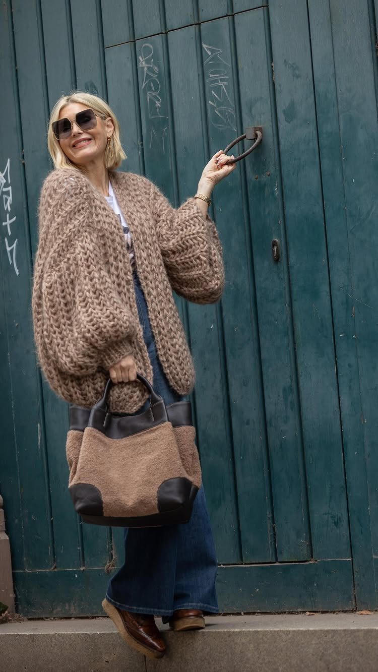 Smiling woman wearing a chunky beige sweater and blue wide-leg jeans standing by a teal door.
