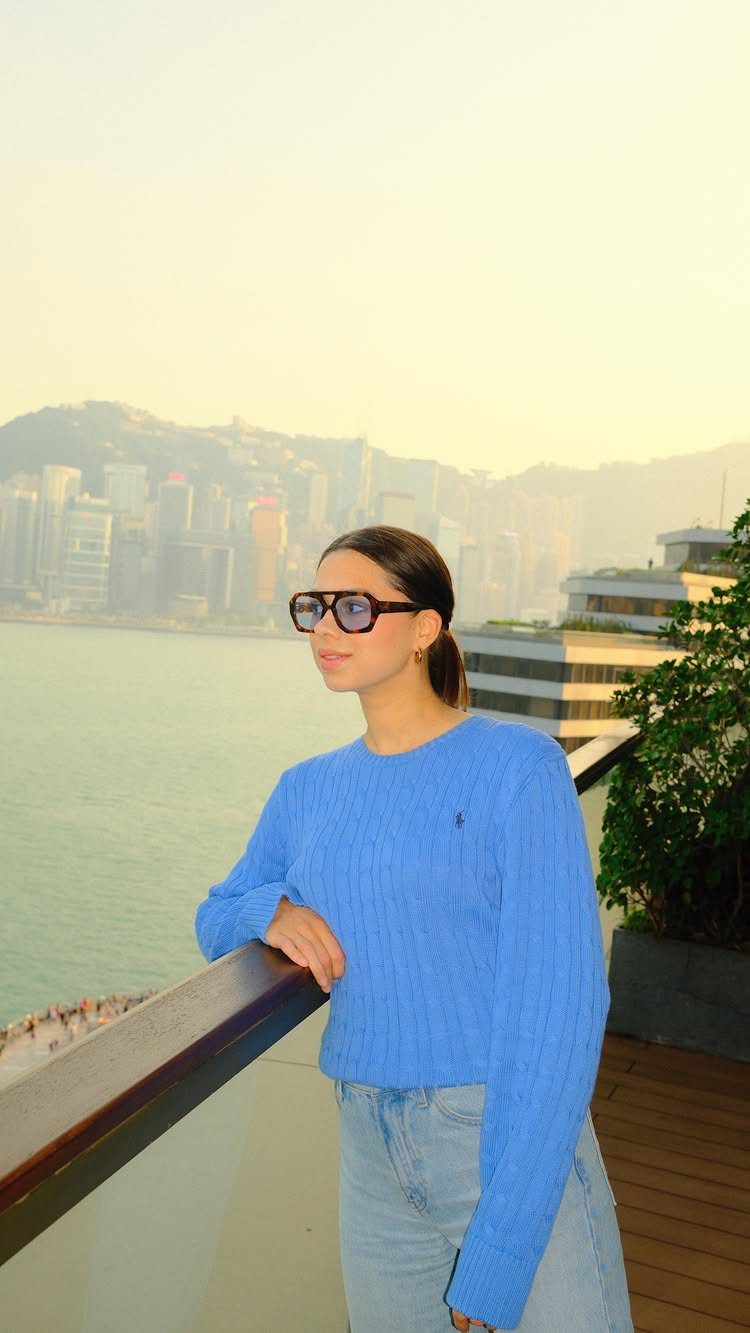 Woman in a blue sweater and light blue jeans standing on a balcony overlooking a city.