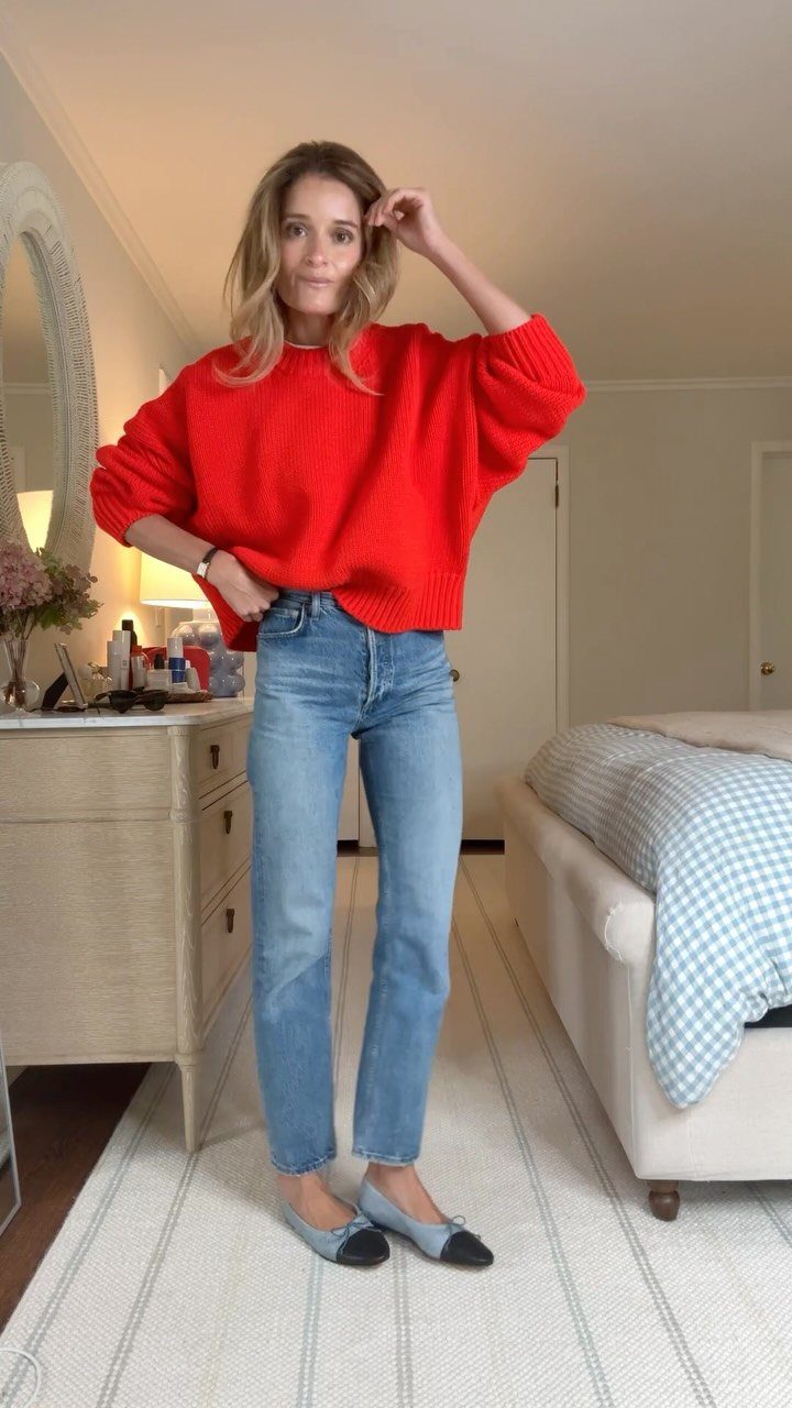 A woman posing in a bright red sweater and light blue denim jeans with grey flats.