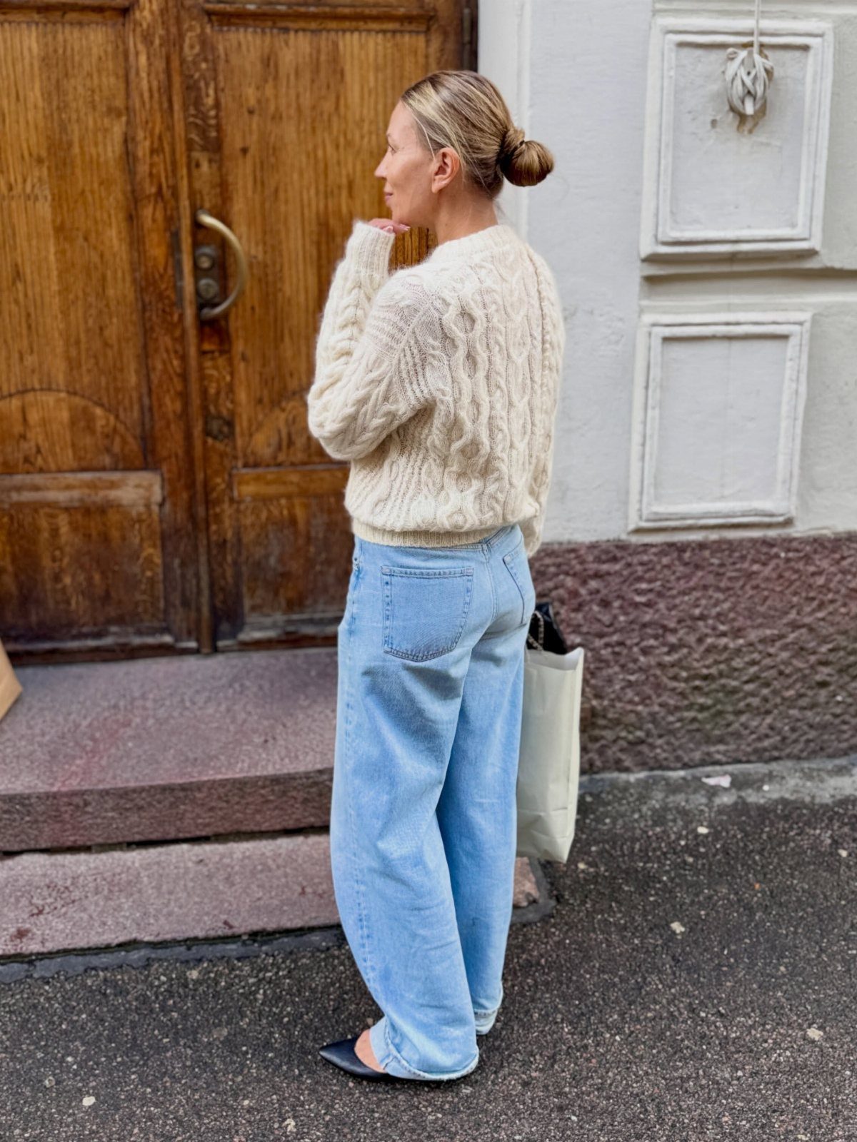 Woman wearing a cream cable-knit sweater and light blue wide-leg jeans standing by a wooden door.