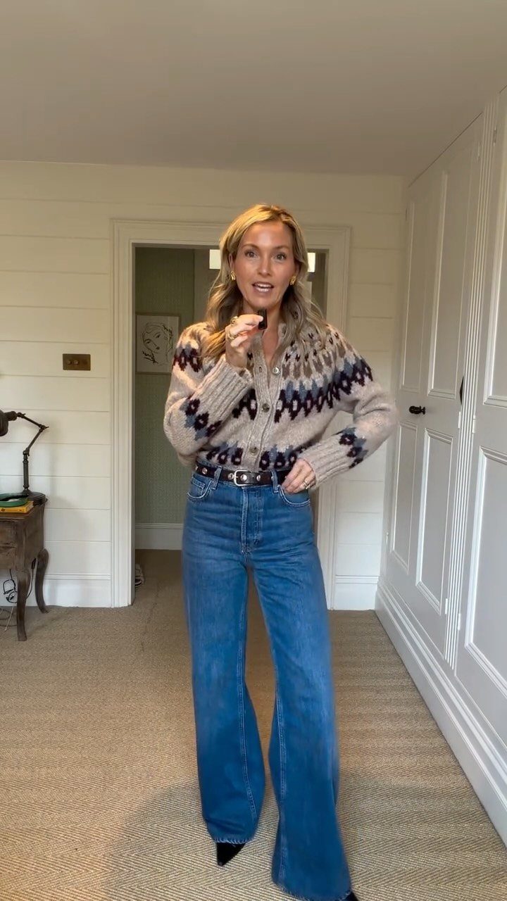 A woman posing in a patterned beige sweater and blue flared denim jeans indoors.