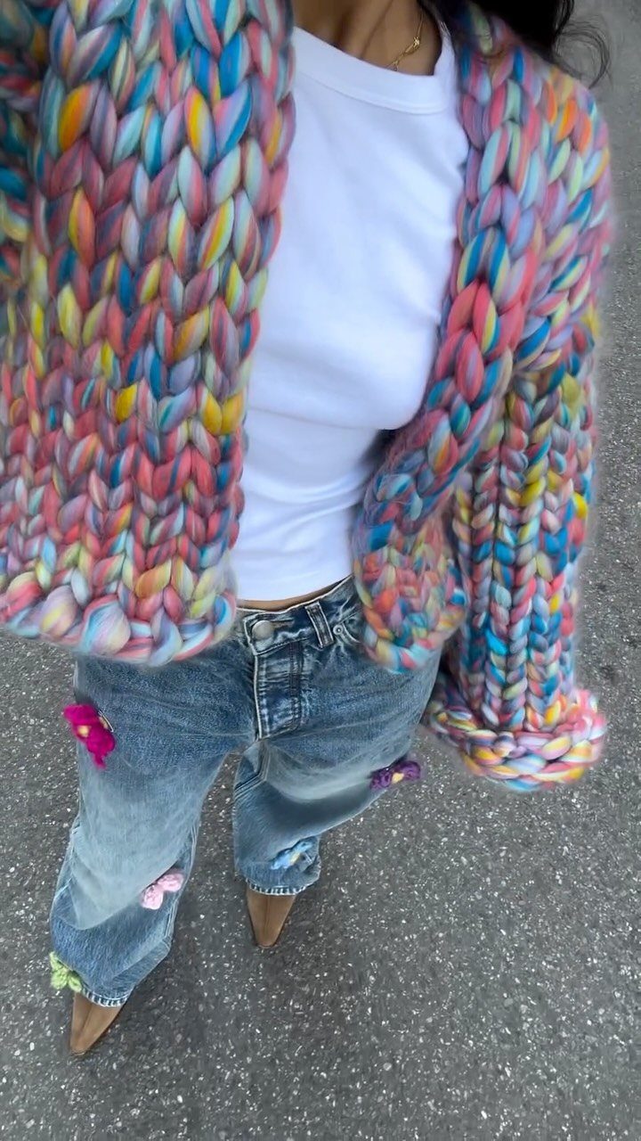 A person wearing a colorful chunky knit sweater and blue straight-leg jeans with floral patches.