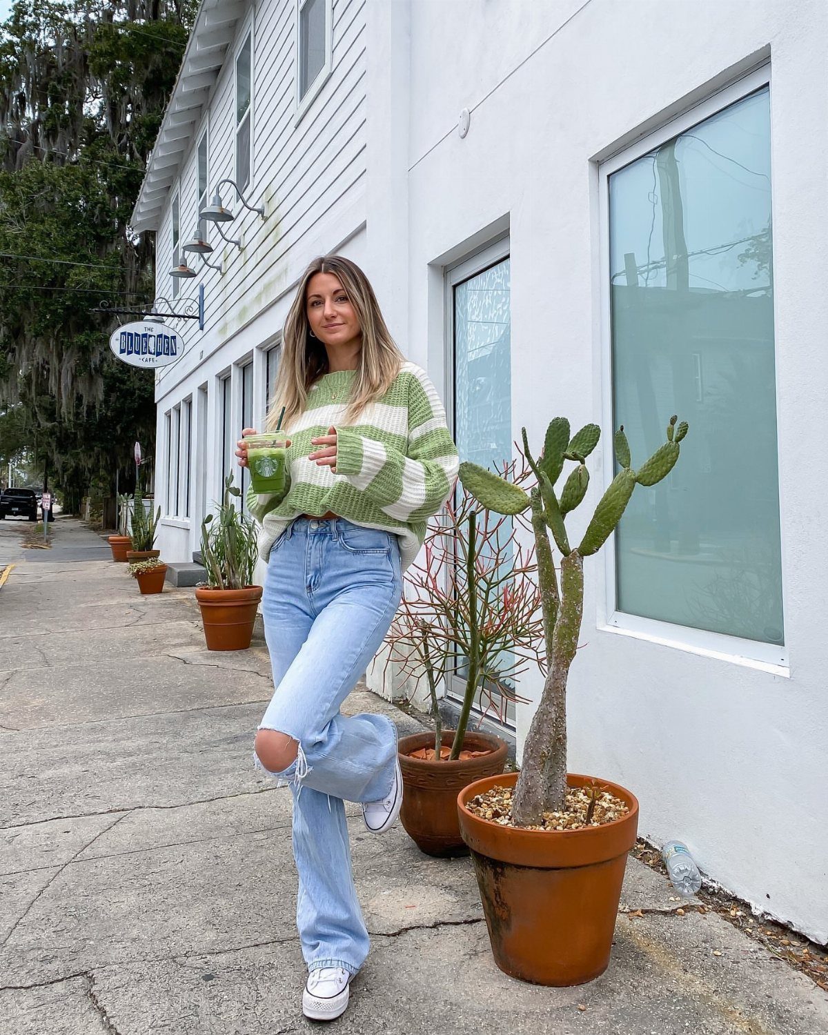 Woman in a striped green sweater and light blue jeans standing on a sidewalk by potted plants.