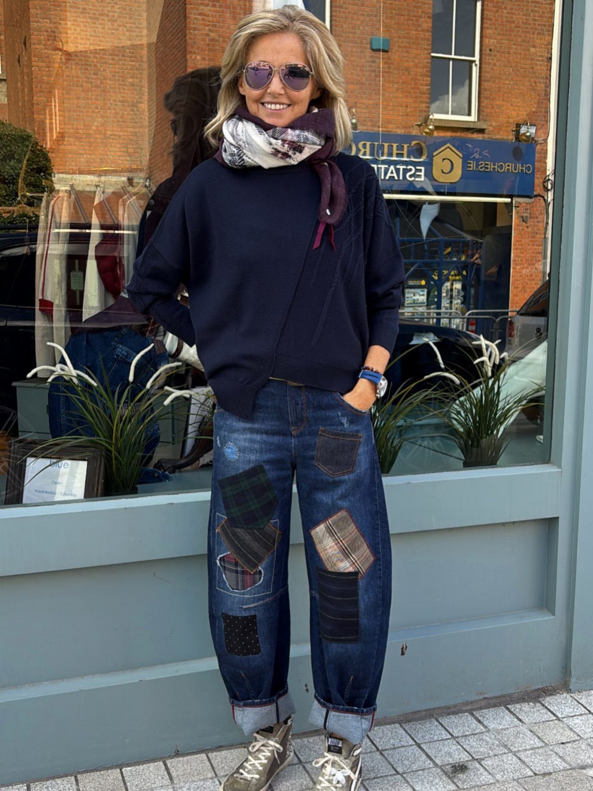 A smiling woman wearing a navy sweater, patterned scarf, and patchwork jeans standing outdoors.