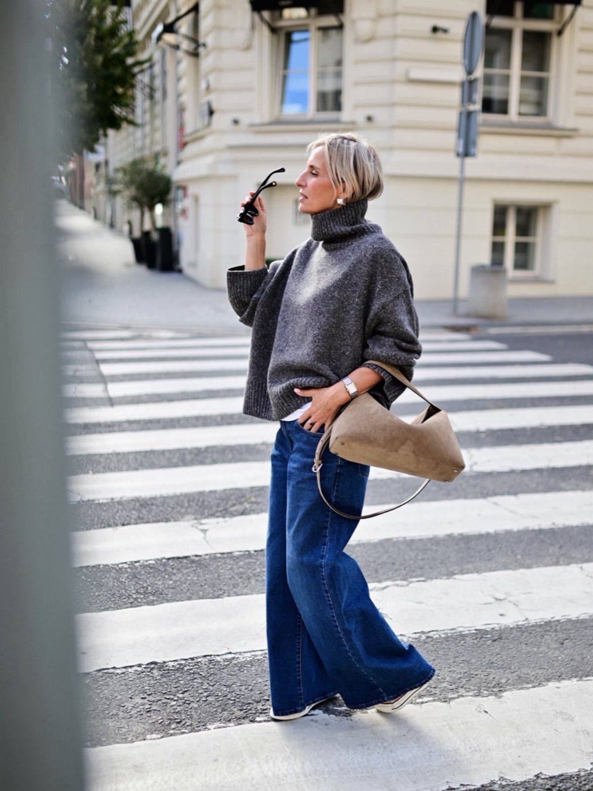 A woman walking in a city wearing a grey turtleneck sweater and blue wide-leg jeans.