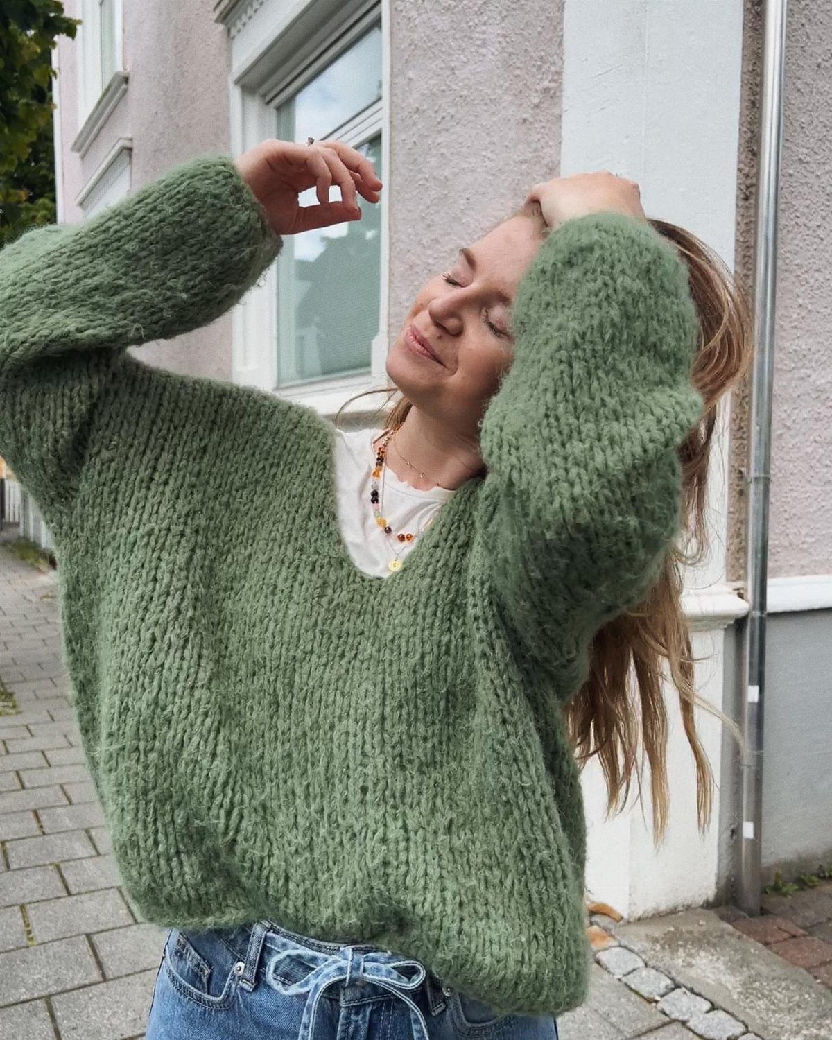 A woman in a fuzzy green v-neck sweater and blue jeans posing outdoors.