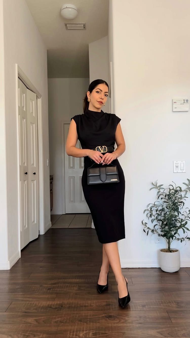 Woman in a sleeveless black midi dress posing in a hallway with a black handbag.