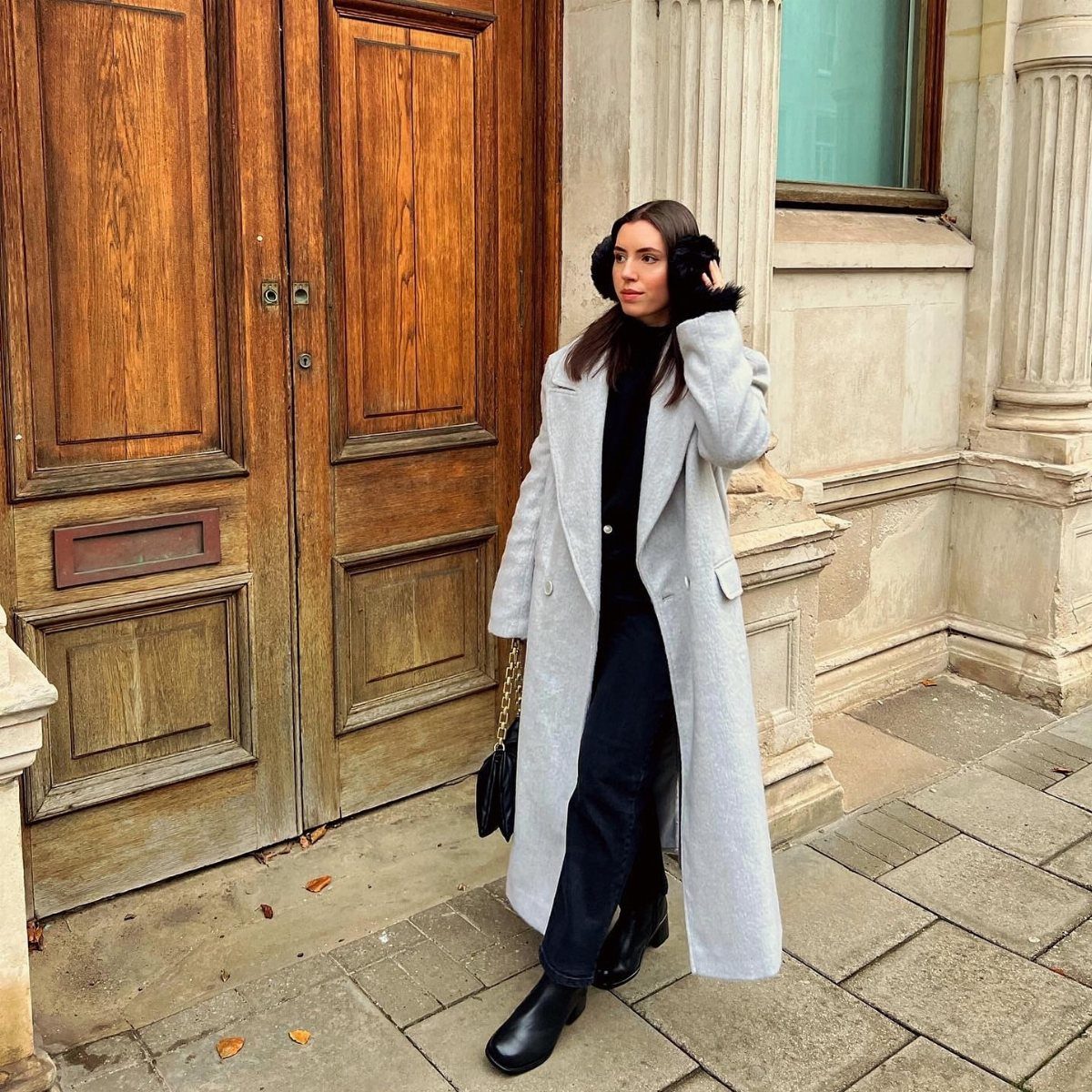 Woman wearing a long grey coat and black earmuffs standing by large wooden doors.