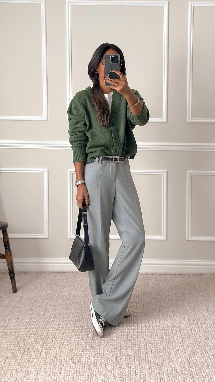 A woman posing in a mirror wearing a green cardigan and grey tailored trousers.