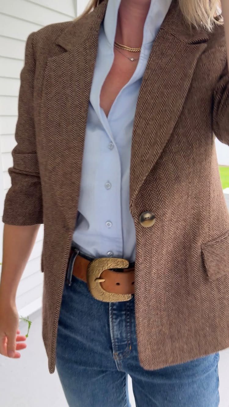 A woman wearing a brown textured blazer, light blue shirt, and blue jeans with a gold buckle belt.
