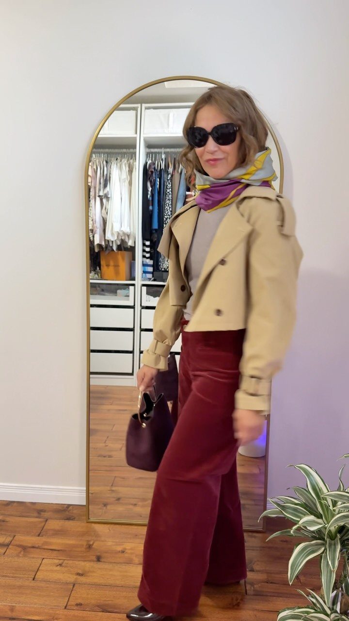 A woman wearing a tan trench coat and burgundy wide-leg trousers posing in front of a mirror.