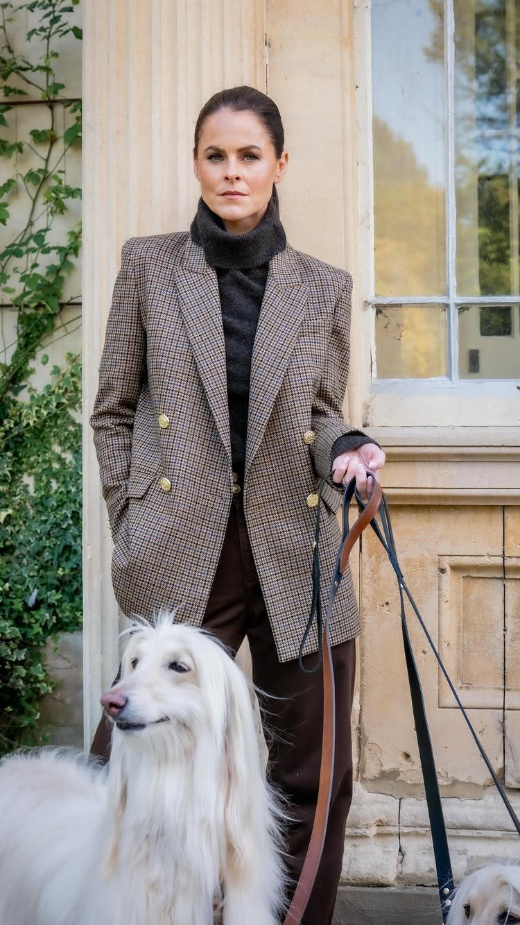 A woman in a patterned blazer and turtleneck standing with a white long-haired dog.