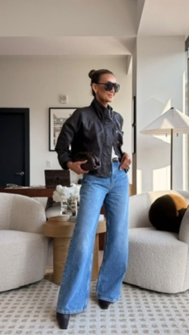 A woman posing in a black leather jacket and wide-leg blue jeans indoors.