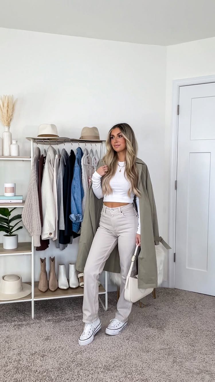 A woman posing in a white top, beige pants, and an olive green trench coat.