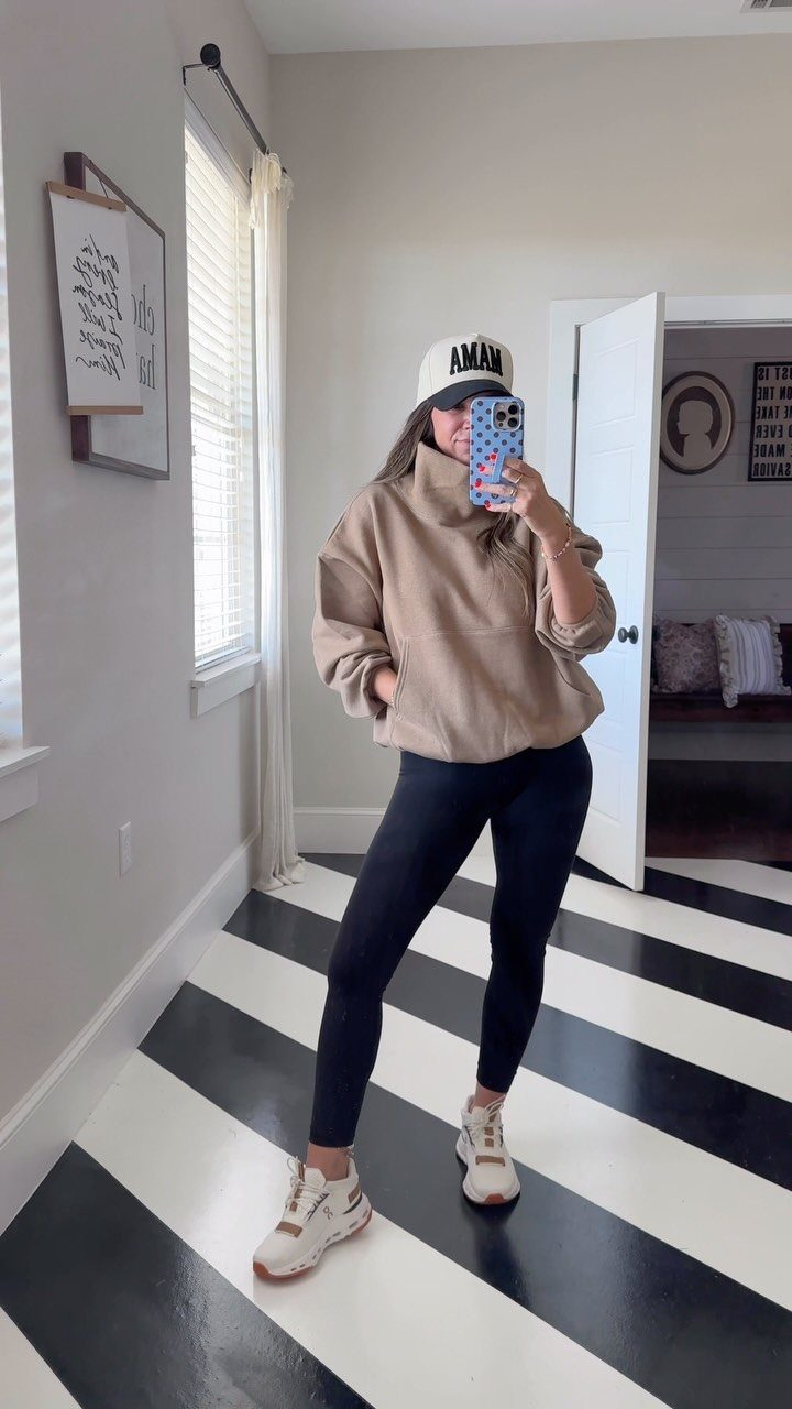 Woman in tan sweatshirt, black leggings, and white sneakers posing for a mirror selfie.