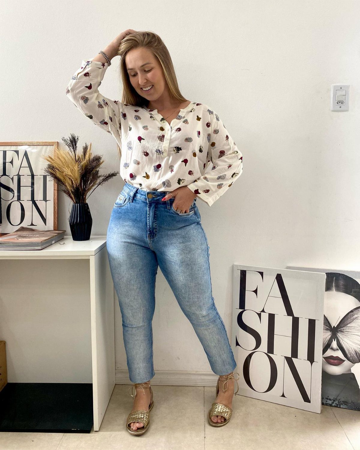 A woman wearing a patterned white blouse and light blue jeans standing in a bright room.