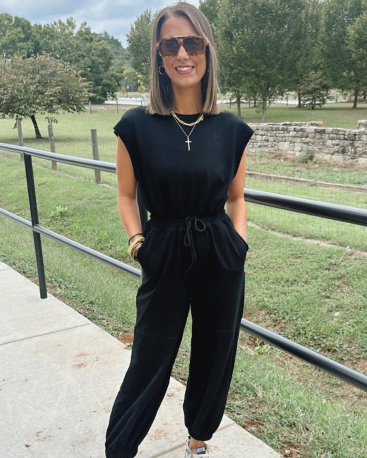 A smiling woman wearing a black sleeveless jumpsuit and sunglasses standing outdoors by a railing.