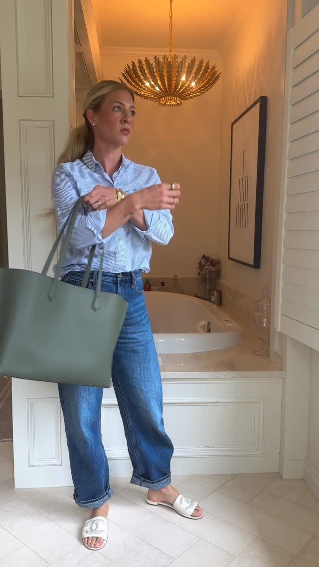 Blonde woman wearing a blue striped button-down shirt, blue jeans, and white sandals holding a large green tote.