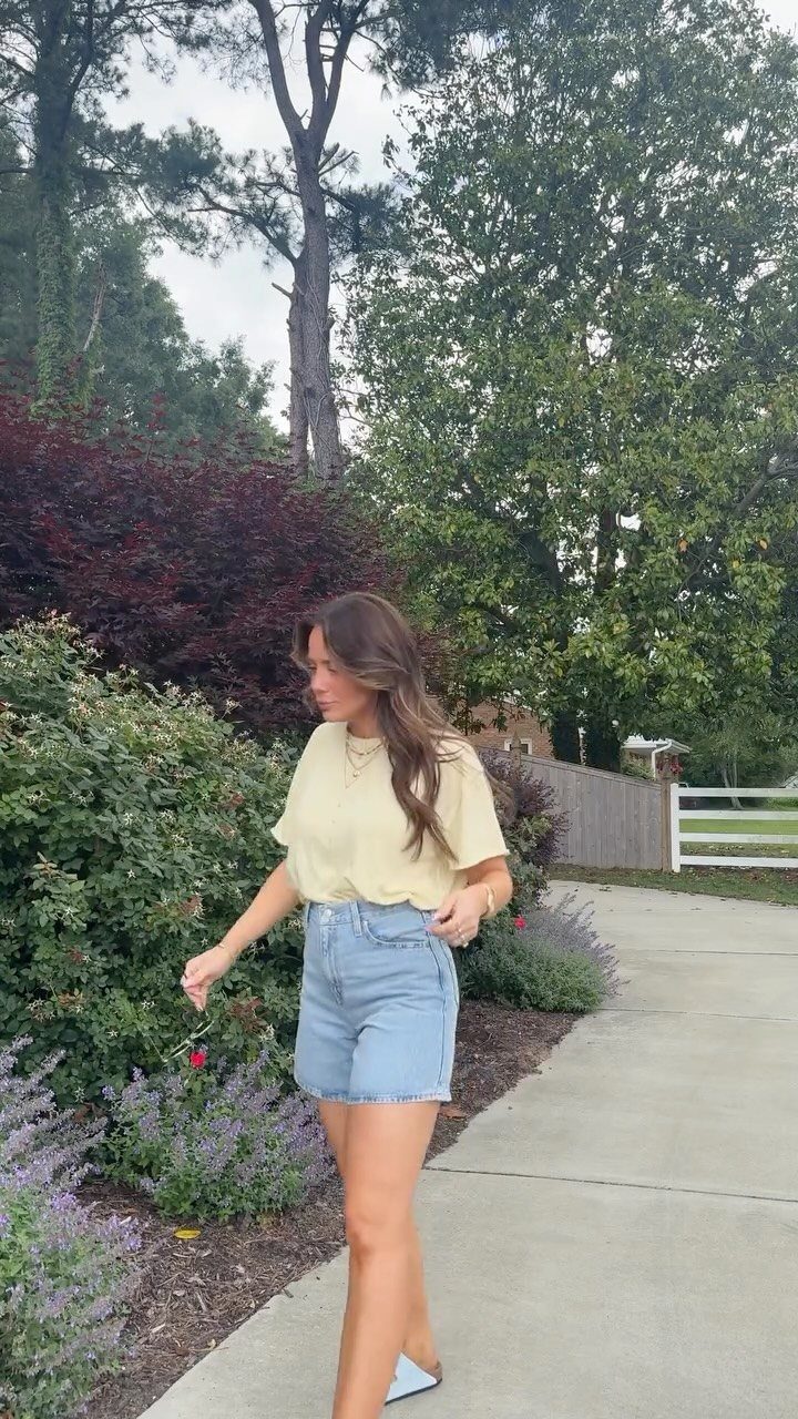 A woman wearing a yellow t-shirt and denim shorts walking on a path near green bushes.