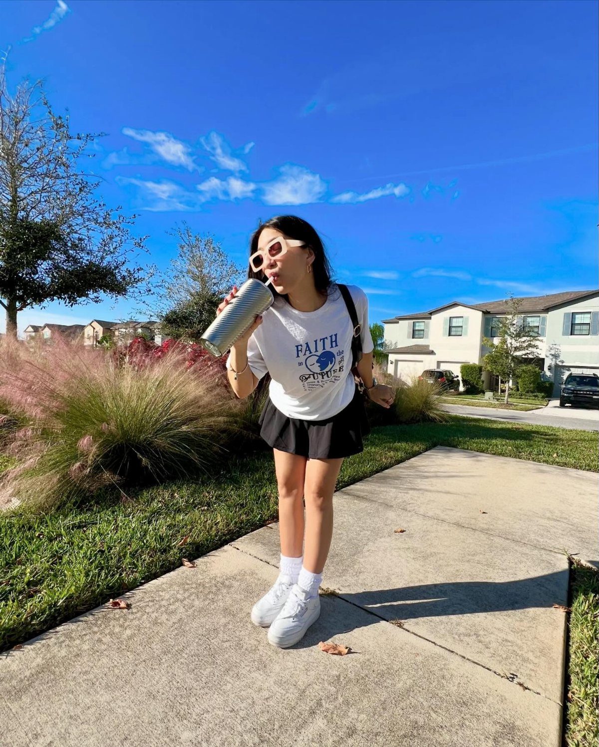 A woman in a white t-shirt and black skirt standing on a sunny sidewalk.