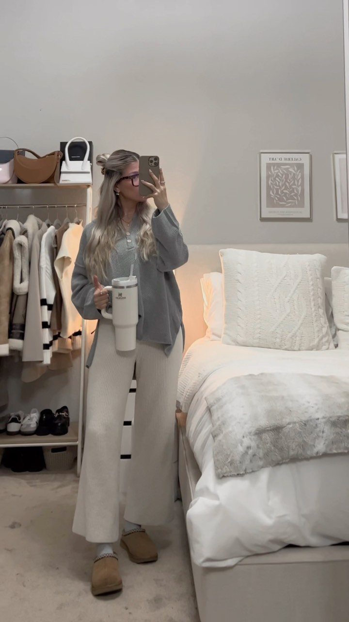 Woman in blue top and white ribbed trousers holding a tumbler in a bedroom.