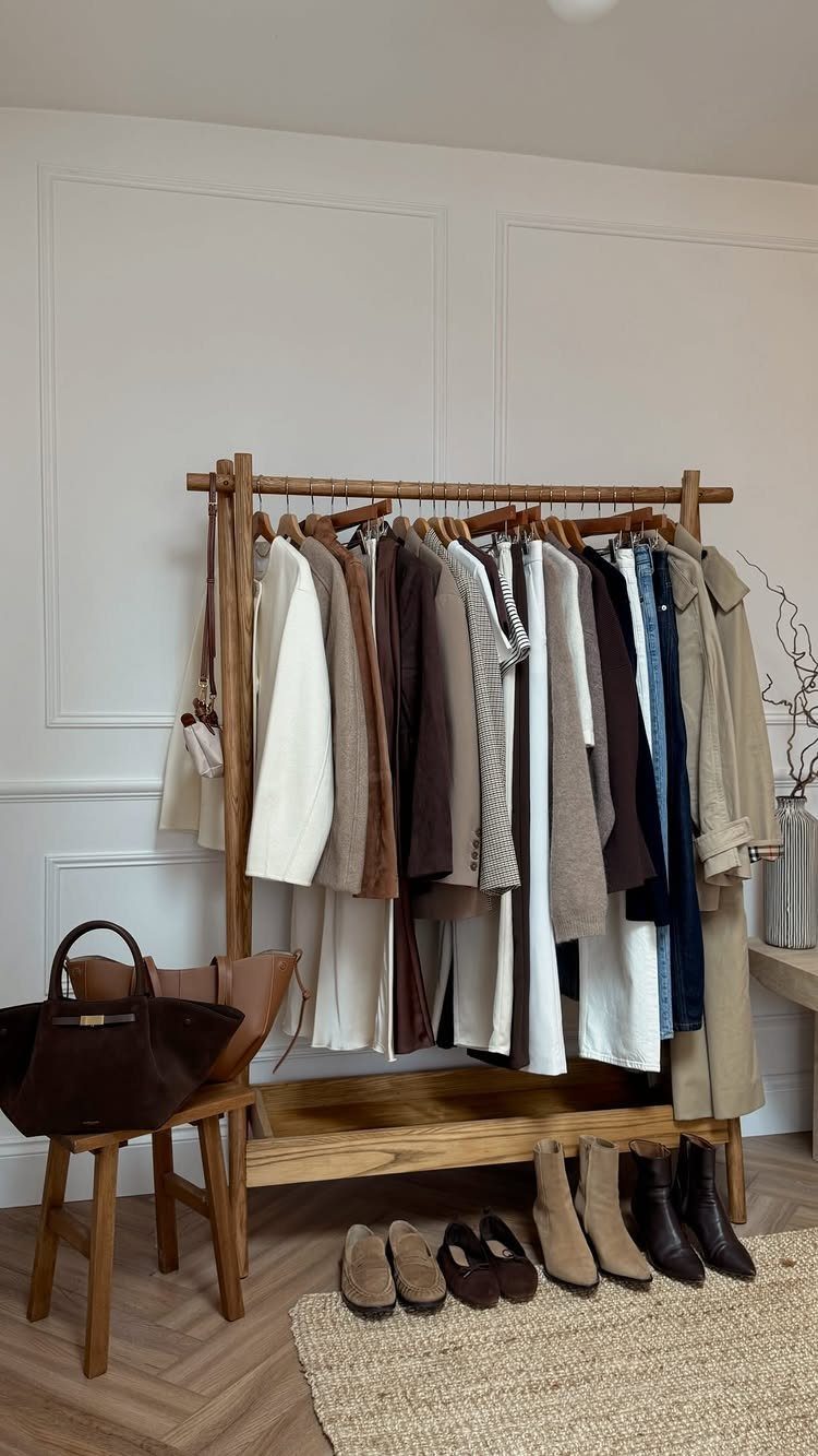A wooden clothing rack filled with neutral and warm toned garments and boots on the floor.