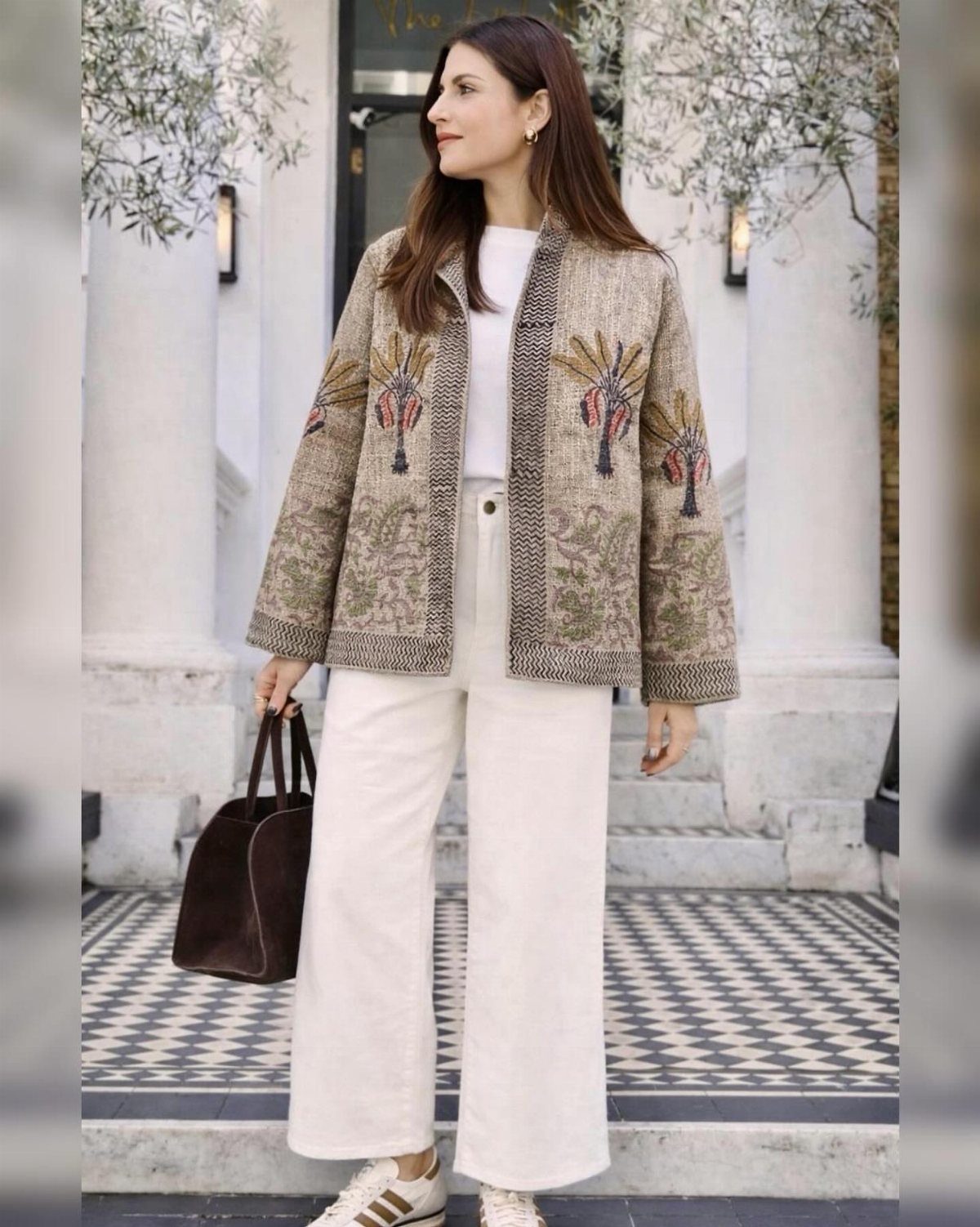 Woman wearing a patterned beige jacket, white top, and wide-leg trousers while holding a brown handbag.