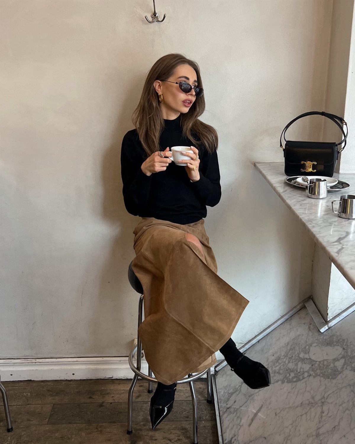 Woman in a black turtleneck, tan midi skirt, and black high heels sitting in a cafe.