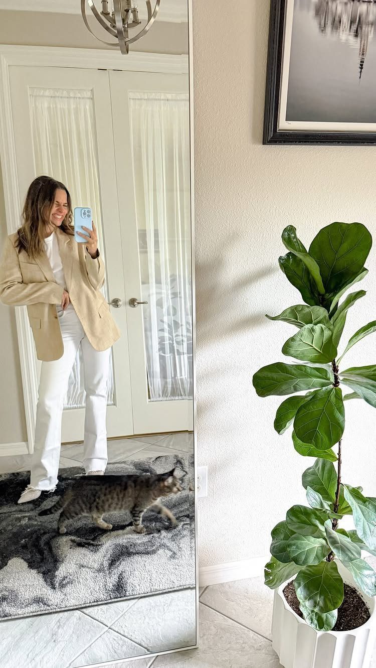 Woman in white jeans and a beige blazer taking a mirror selfie indoors.