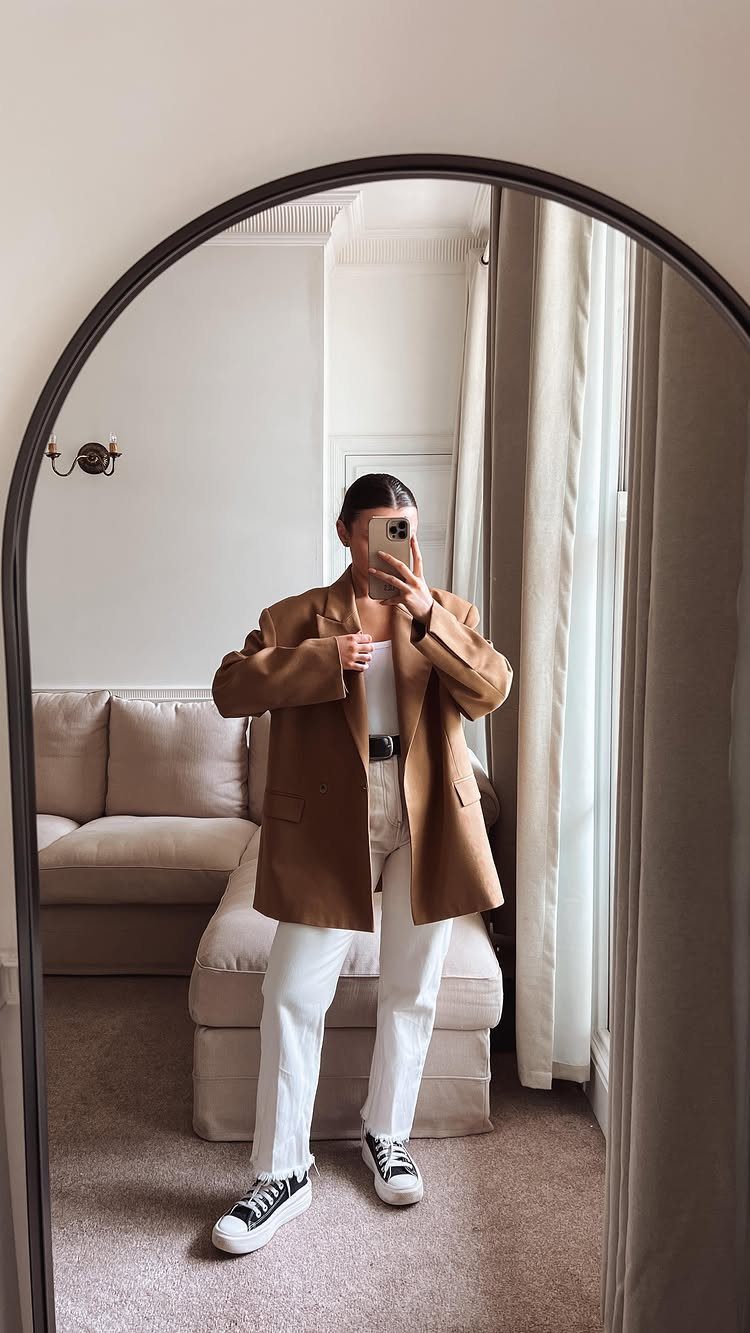 Person in white jeans, white top, brown blazer, and black sneakers taking a mirror selfie.