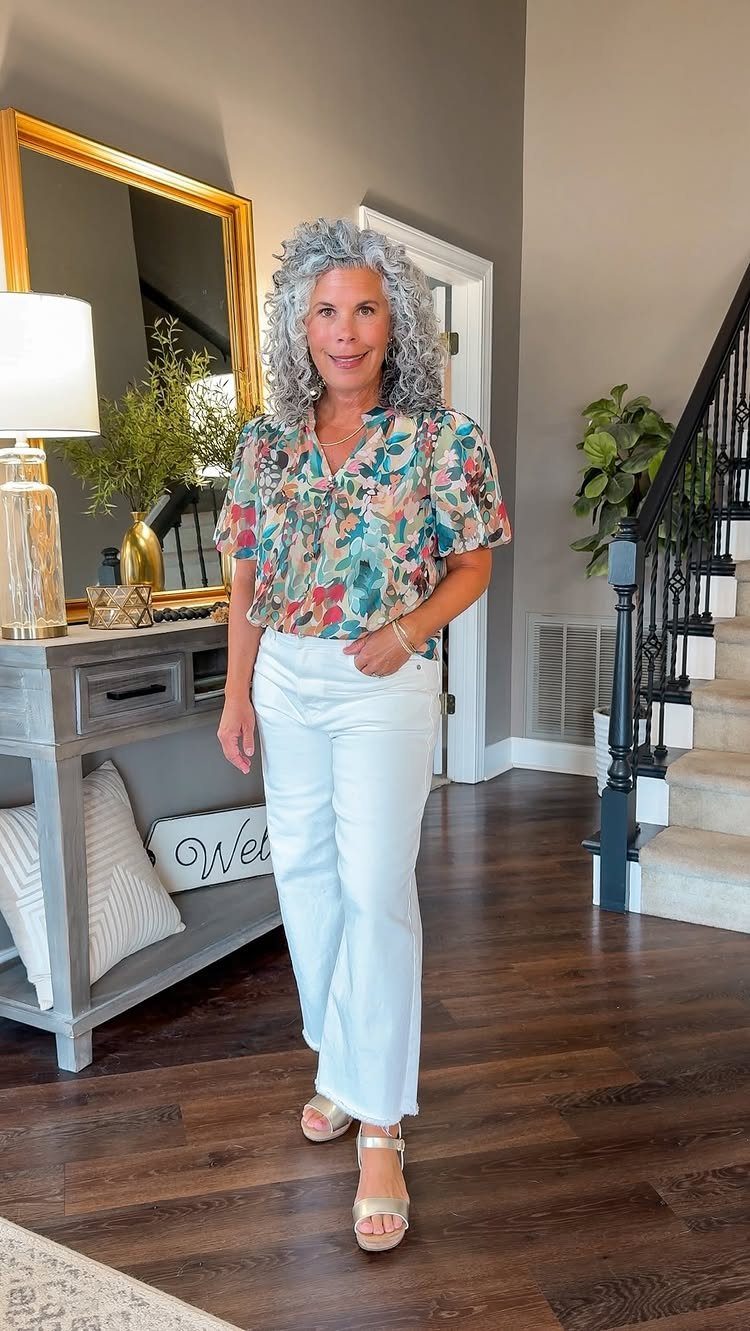 A woman posing in a colorful floral blouse and white wide-leg jeans indoors.