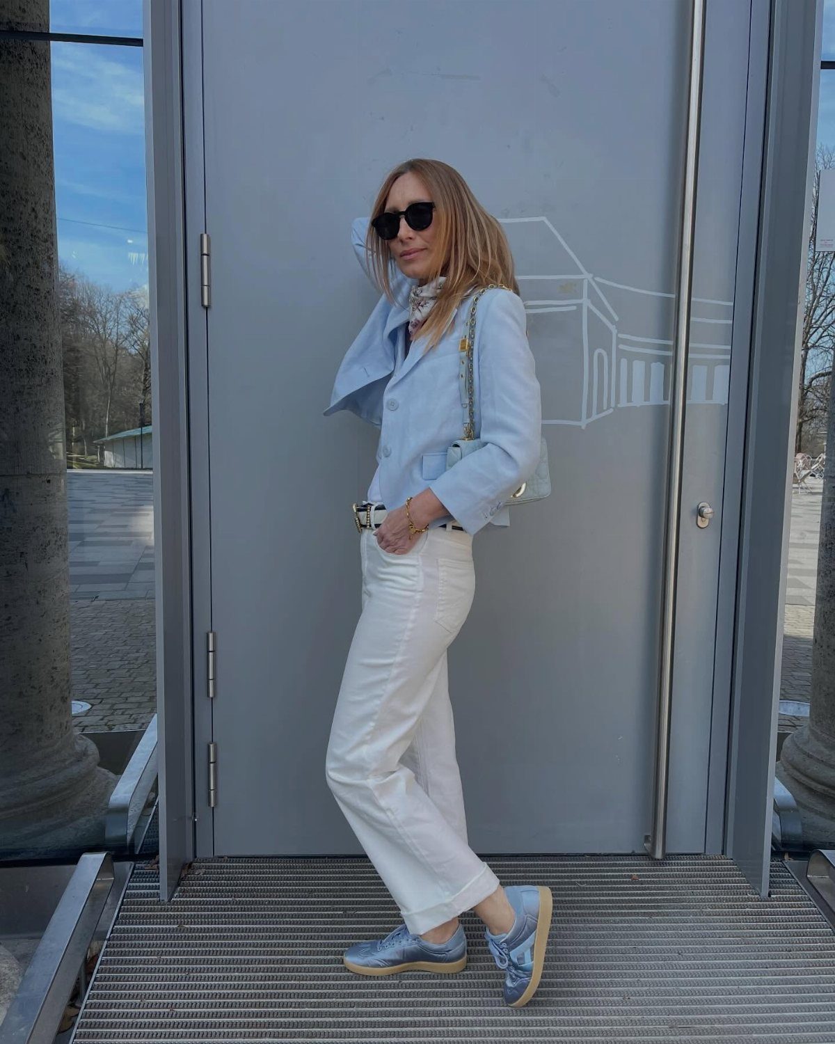 Woman in white jeans, light blue blazer, and blue satin sneakers posing outdoors.