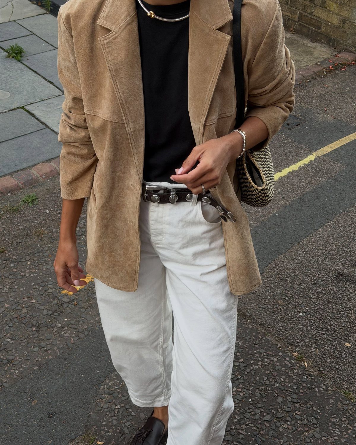 A person wearing white jeans, a black t-shirt, and a tan suede blazer outdoors.