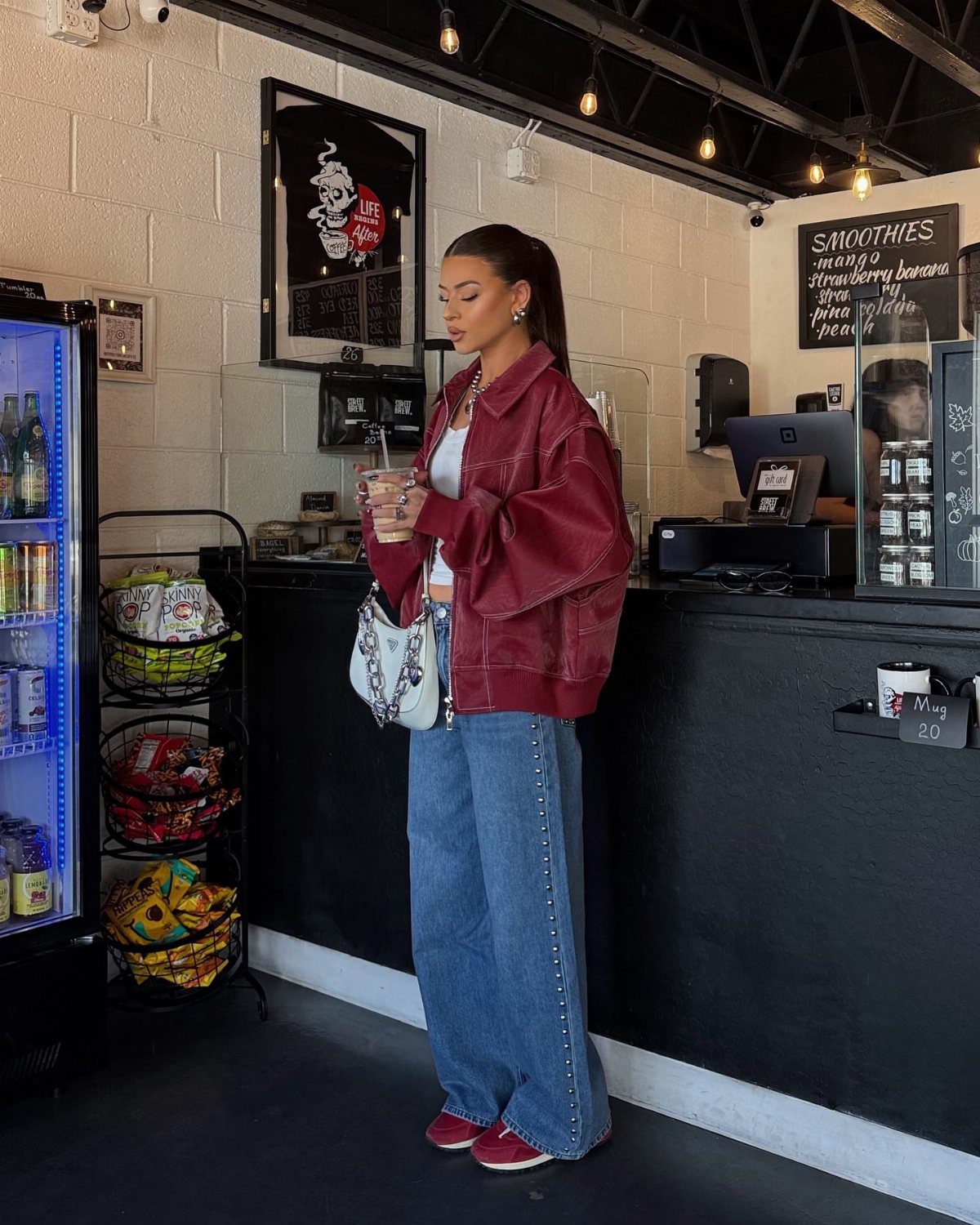 Woman wearing a burgundy leather jacket and blue wide-leg jeans standing in a cafe.