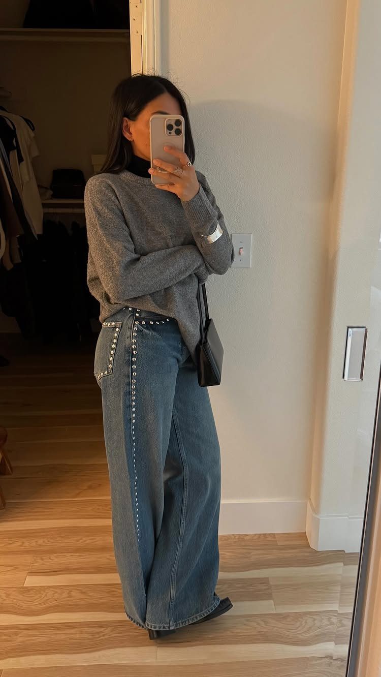 A woman posing in a mirror wearing a grey sweater and wide leg denim jeans.