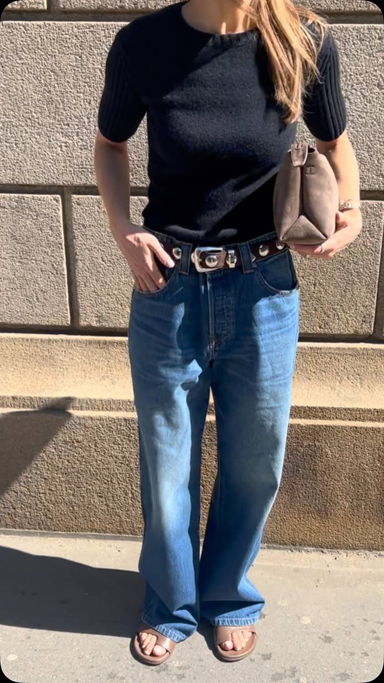 A person wearing a black top and blue wide leg jeans standing against a stone wall.
