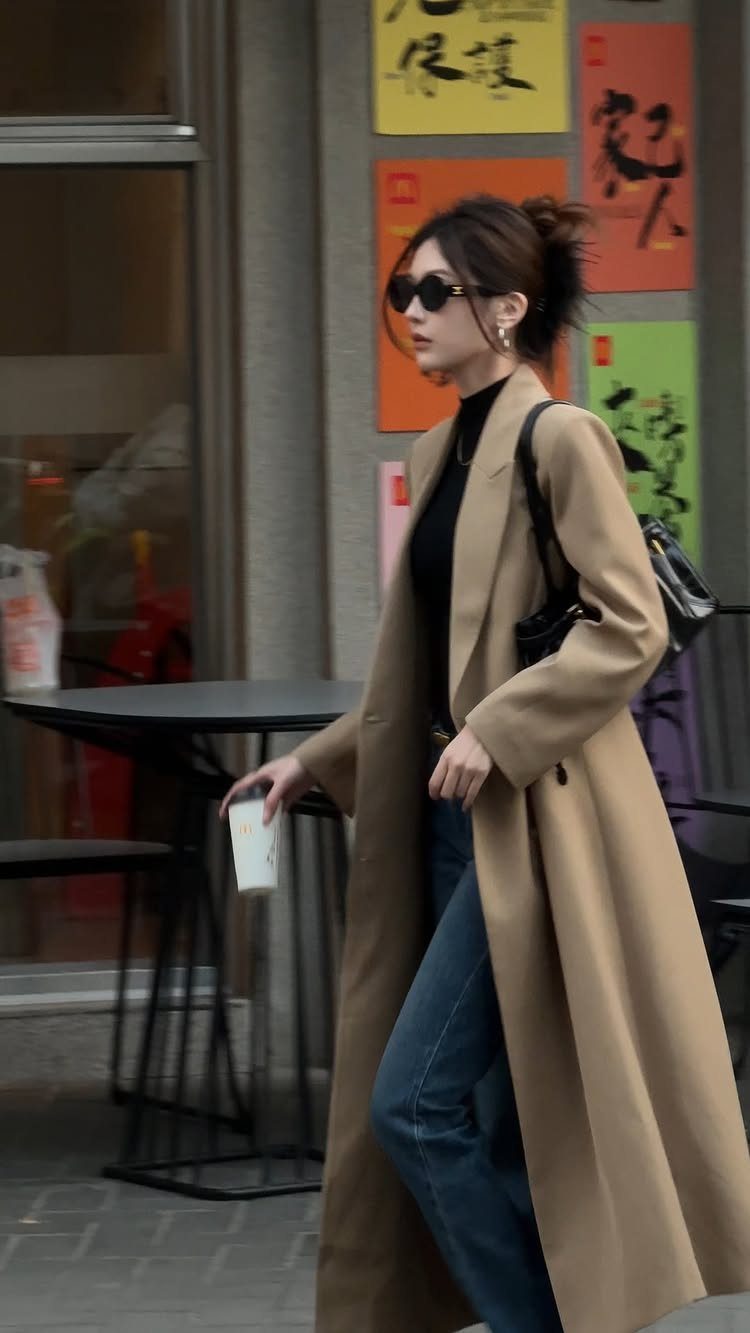 A woman in a tan coat and blue wide-leg jeans walking past a cafe.